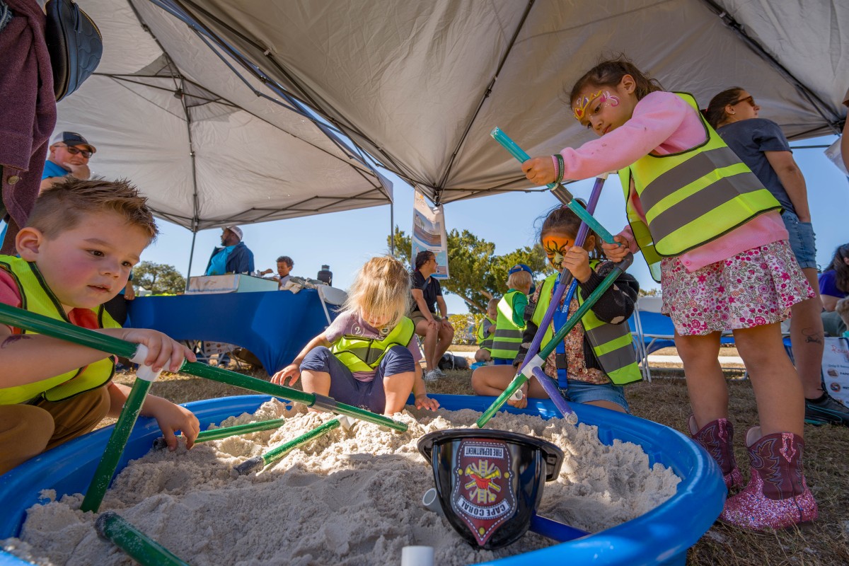 Children in yellow safety vests dig with toy tools in a sand-filled pool at the Waterways Festival.